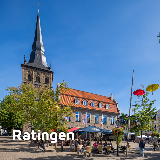 Ratinger Marktplatz im Sommer. Im Hintergrund ist die Kirche und das Brauhaus zu sehen, davor sitzen Menschen. Über den Straßen hängen bunte Regenschirme.