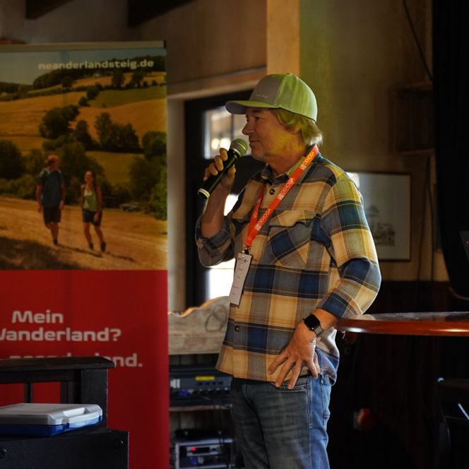 Ein Mann steht auf einer Bühne und spricht ins Mikrofon, im Hintergrund ist ein Roll-Up mit Wandernden zu sehen. 