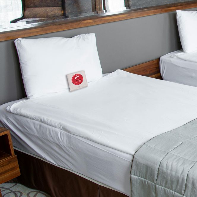 Vitalbox auf einem Bett im Hotelzimmer mit Siegel vom neanderland STEIG Qualitätsbetrieb Das Bild zeigt zwei Betten in einem Hotelzimmer. Auf einem Bett liegt eine kleine Vitalbox mit dem Siegel vom Qualitätsnetzwerk des neanderland STEIGs.