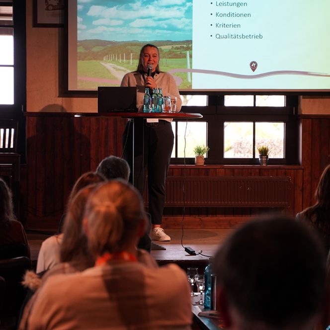 Eine Frau steht auf einer Bühne und trägt einen Vortrag vor, im Vordergrund ist das interessierte Publikum zu sehen. 