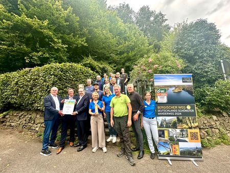 Ein Gruppenfoto von mehreren Personen stehen vor einer grünen Hecke, rechts im Bild ein Werbebanner mit der Aufschrift Bergischer Weg - Deutschlands schönster Wanderweg 2024.