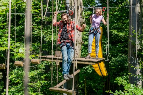 Mann balanciert auf waagerechten Baumstämmen auf einem Parcours im Waldkletterpark Velbert-Langenberg mit Frau im Hintergrund.