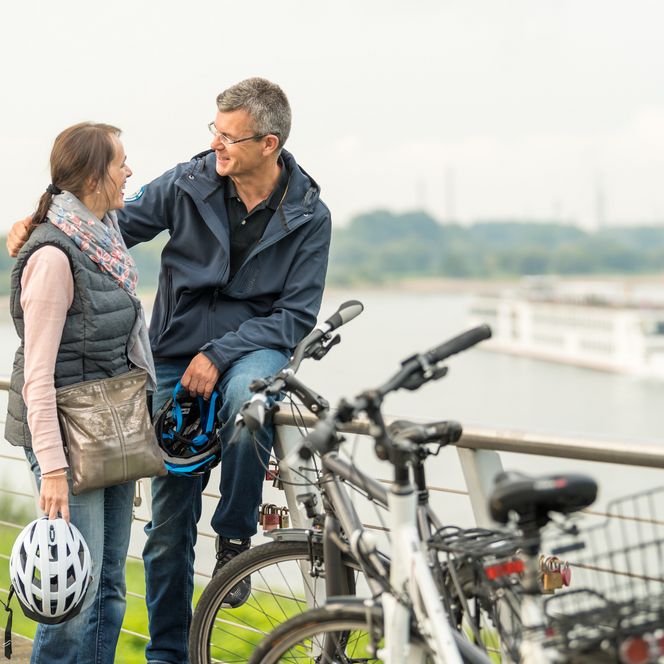 Mann umarmt Frau neben zwei Fahrrädern auf einer Aussichtsplattform am Rhein nahe des NRW-Rheinradweges mit Schiff im Hintergrund