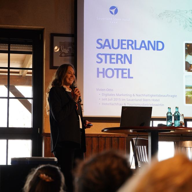 Eine Frau hält eine interessante Rede auf einer Bühne, im Vordergrund ist das Publikum von hinten zu sehen. 