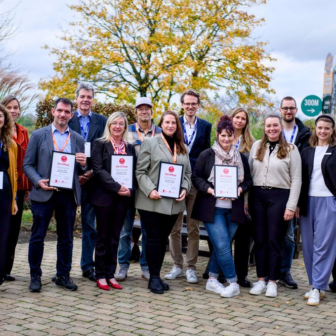 Gruppenfoto von 14 Personen, die freundlich in die Kamera schauen, im Hintergrund steht ein großer Baum mit vom Herbst gefärbten gelben Blättern. 