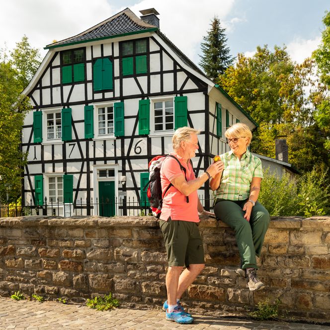 Frau und Mann pausieren an einer Mauer vor einem alten Fachwerkhaus in Haan-Gruiten nahe des neanderland STEIGs