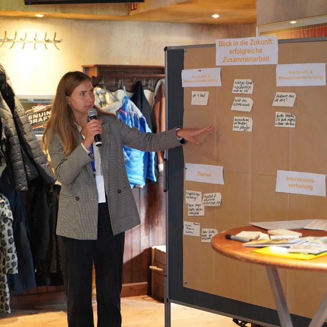 Eine Frau steht neben einer mit Zetteln beklebten Stellwand und spricht zum Publikum.