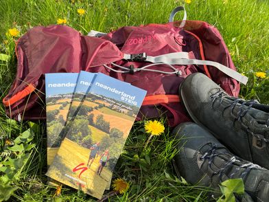Immagine di nuovo neanderland STEIG Brochure escursionistica adagiata su un prato con zaino e scarponi da trekking