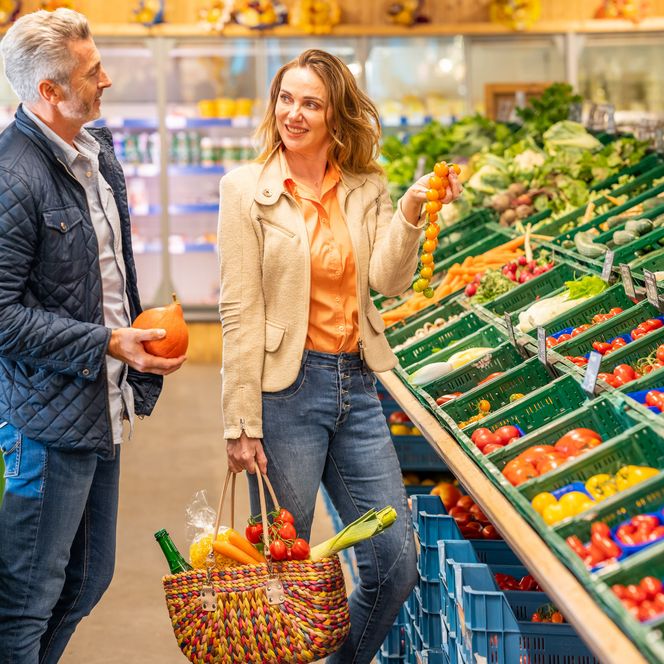 Ein Mann und eine Frau stehen vor einer reich gefüllten Gemüseauslage. Die Frau trägt einen Einkaufskorb, der bereits mit Gemüse gefüllt ist. In der Hand hält sie Tomaten, die sie dem Mann zeigt. Der Mann hält einen Kürbis in der Hand. 