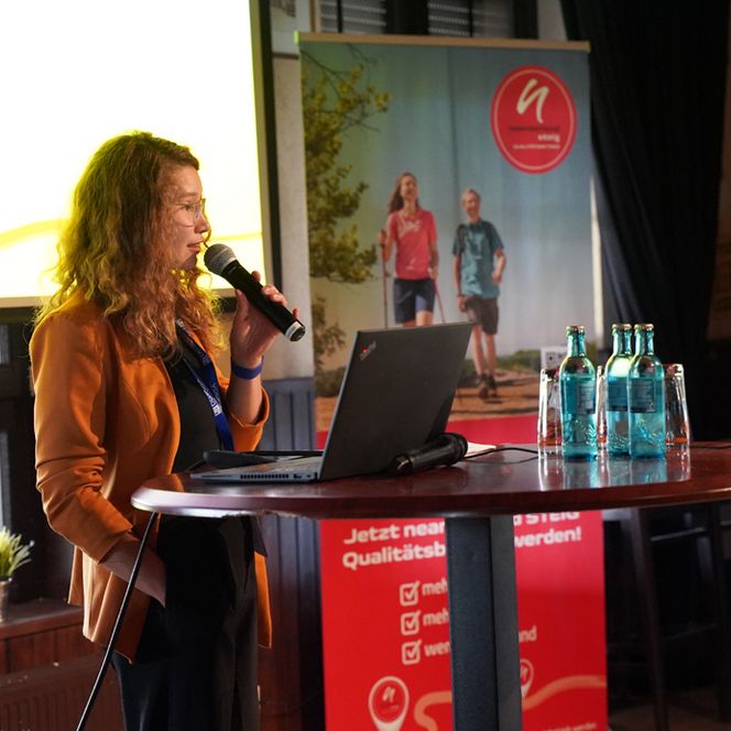 Eine Frau spricht auf einer Bühne ins Mikrofon. Im Hintergrund ist ein Roll-Up mit einem Bild von Wandernden zu sehen. 