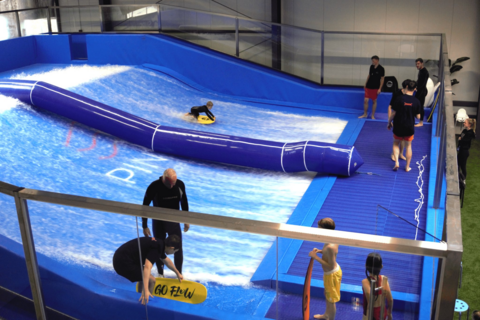 Surfschule mit blauer Indoor-Welle und mehreren Personen, die das Surfen üben.