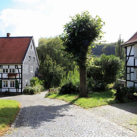 Wejście do Biohof Judt z żółtym znakiem na pierwszym planie i drzewami i krzewami w tle między dwoma domami z muru pruskiego w dolinie Windrath w Velbert-Nordrath