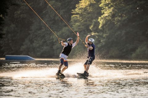 Zwei Wakeboarder geben sich lachend ein High-Five auf der Fahrt mit Bäumen im Hintergrund