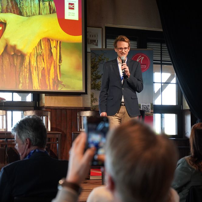 Auf dem Bild sieht man im Hintergrund einen Mann auf einer Bühne stehen der in ein Mikrofon spricht. Im Vordergrund sieht man das interessierte Publikum.