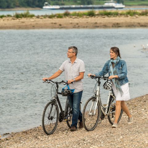 Twee volwassenen lopen met hun twee fietsen langs de Rijn bij het NRW Rijnfietspad