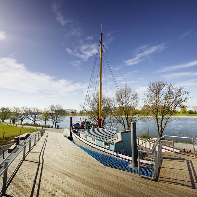 Außenansicht des Aalfischerei-Museums mit Aalschokker "Fiat Voluntas" an einem hölzernen Steg im Vordergrund und dem Rhein im Hintergrund