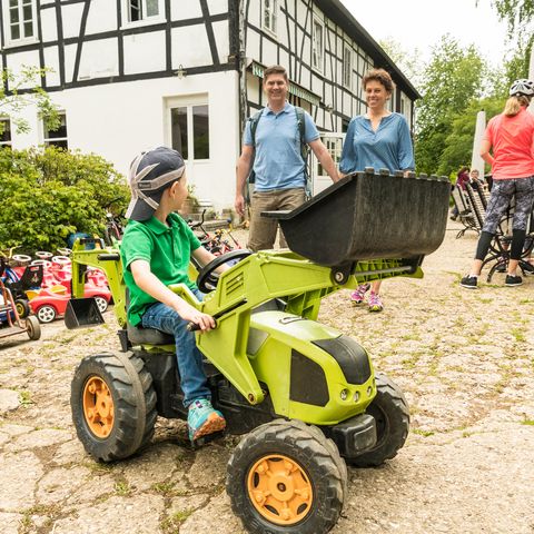 Junge sitzt auf einem grünen Trettraktor draußen auf dem Hof zur Hellen und schaut zu den Eltern im Hintergrund