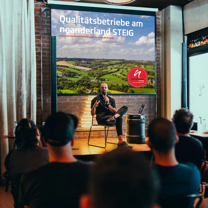 Schulung mit Präsentation und Siegel vom neanderland STEIG Qualitätsbetrieb Das Bild zeigt eine Schulungssituation, in der ein Mann vorne auf der Bühne vor einer Präsentation zum neanderland STEIG dem sitzenden Publikum etwas erklärt.
