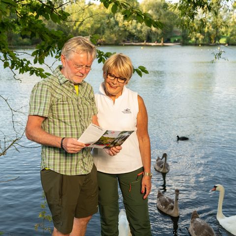 Mujer y hombre leen folletos de senderismo del STEIG neanderland en la orilla con cisnes en el estanque Abtskücher en Heiligenhaus