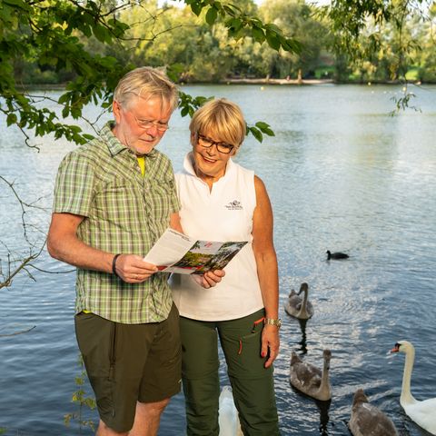 Frau und Mann lesen Wanderflyer des neanderland STEIGs am Ufer mit Schwänen am Abtskücher Teich in Heiligenhaus