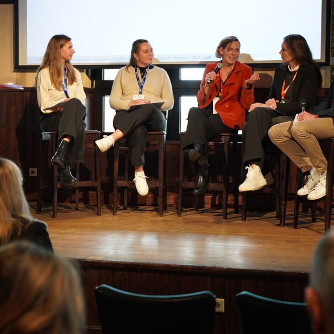 Eine Gruppe von 5 Personen führen auf der Bühne eine Diskussion, im Vordergrund ist das interessierte Publikum zu sehen. 