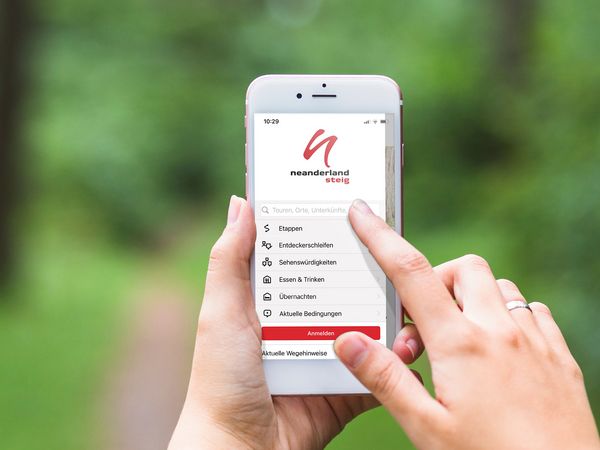 Mock-Up der neanderland STEIG Wander-App (weißes Handy zeigt Wander-App auf Bildschirm bedient durch eine Hand vor grünem Hintergrund)