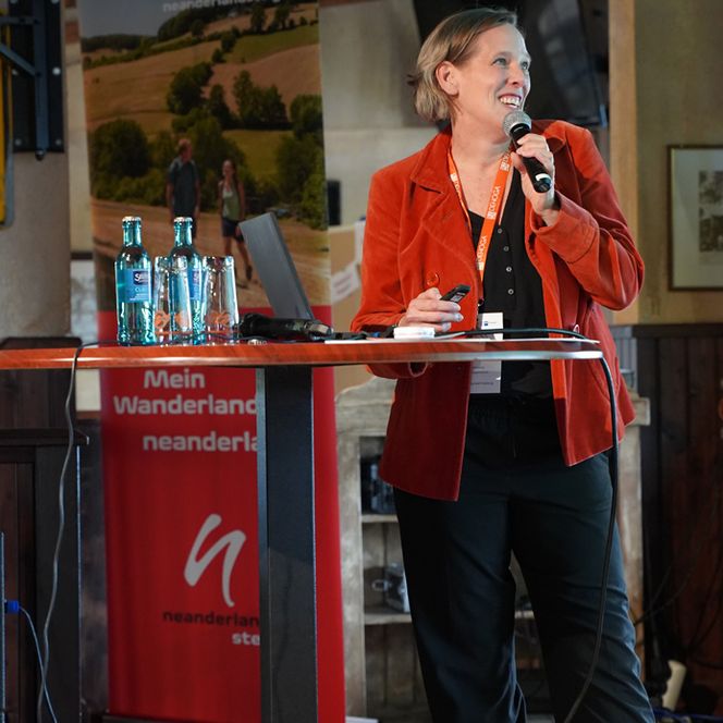 Eine Rednerin lehnt sich lässig an einen Stehtisch an und spricht zum Publikum.