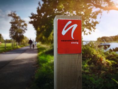 Im Vordergrund die rote Wegemarkierung vom neanderland STEIG, im Hintergrund verschwommen ist links der Wanderweg und rechts der Rhein zu erkennen