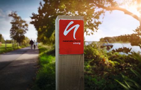 Im Vordergrund die rote Wegemarkierung vom neanderland STEIG, im Hintergrund verschwommen ist links der Wanderweg und rechts der Rhein zu erkennen